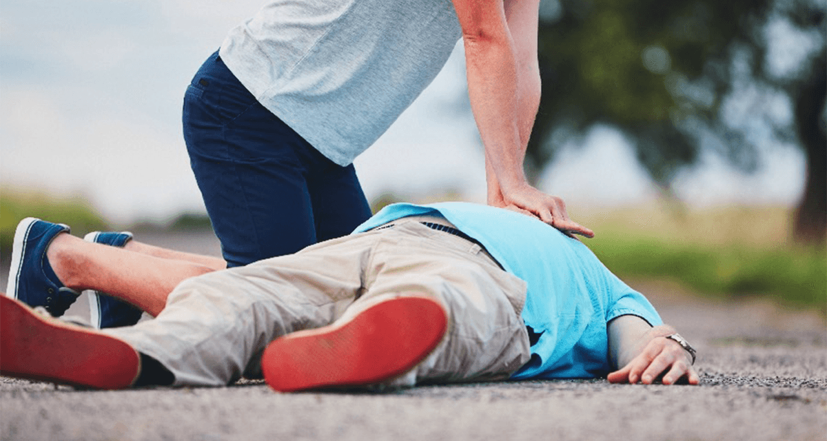 A person in a grey shirt is leaning over a person in a teal shirt who is laying on the ground, and is performing chest compressions as part of CPR
