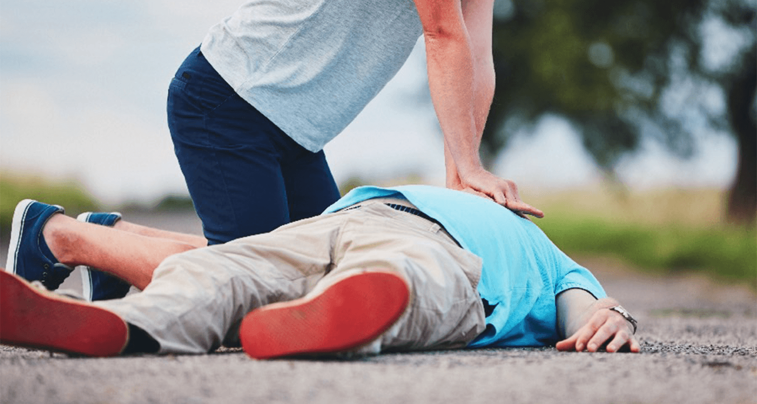 A person in a grey shirt is leaning over a person in a teal shirt who is laying on the ground, and is performing chest compressions as part of CPR